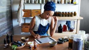 person working at a desk with a tablet computer surrounded by jars and shelves of herbs and tinctures