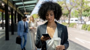 Black business woman at the bus stop holding her take out coffee and looking at her smartphone