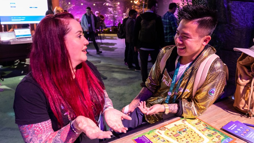 Two Dreamforce attendees (a woman in a black shirt and a man in a gold Trailblazer jacket) sit at a table and have a fun discussion