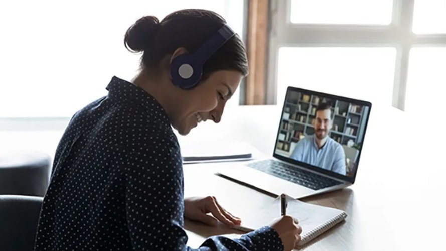 A woman sitting at a desk and taking notes, while on a video call with a colleague.