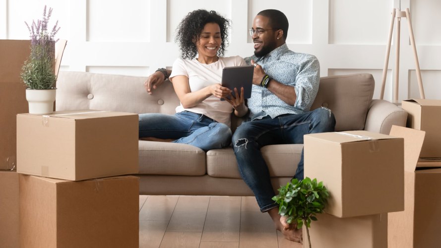 female and male couple sitting on a sofa looking at a tablet while surrounded by moving boxes mortgage lending solutions