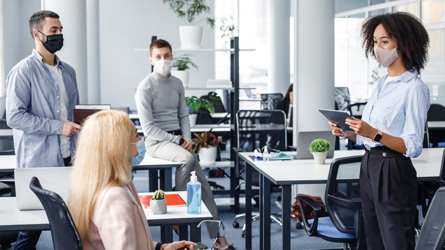 Four coworkers in an office wearing masks and talking, perhaps about change management best practices.