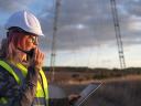 A woman wearing a hard hat views telecom innovations on a tablet near a cell phone tower