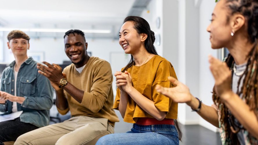 Smiling woman in an office sitting in a circle with fellow employees, who are applauding her / customer experience and profitability