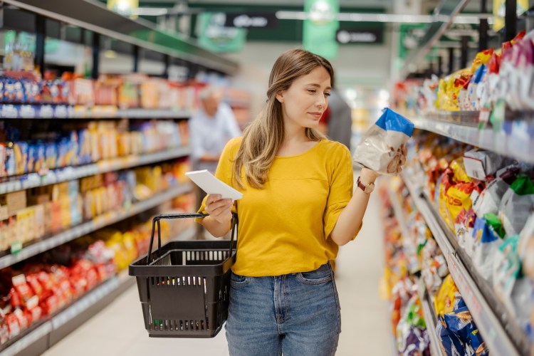A woman reads the label of a food package in the aisle of a grocery store, a common consumer behavior.