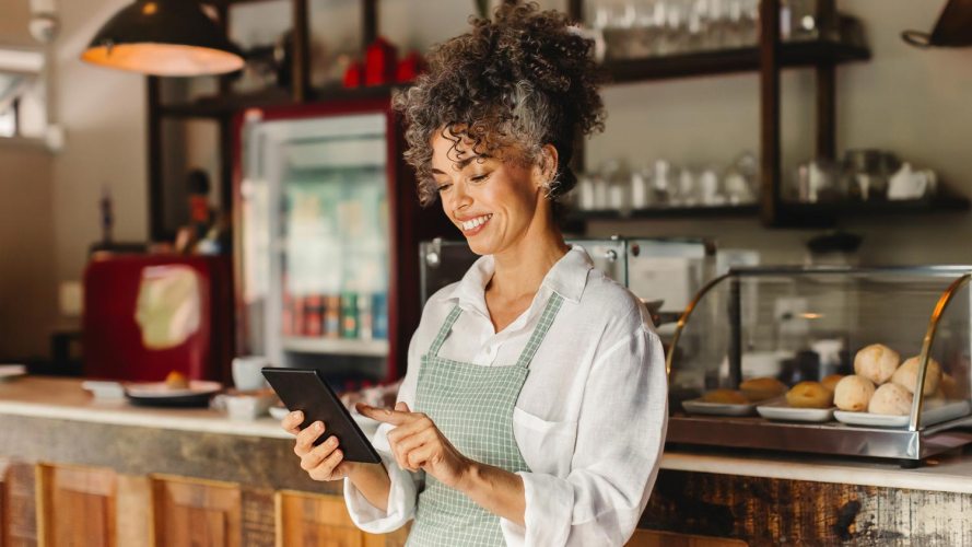 Woman smiling in a bakery, wearing a green apron, holding a tablet and learning more about how automation from the communications industry can help her small business.