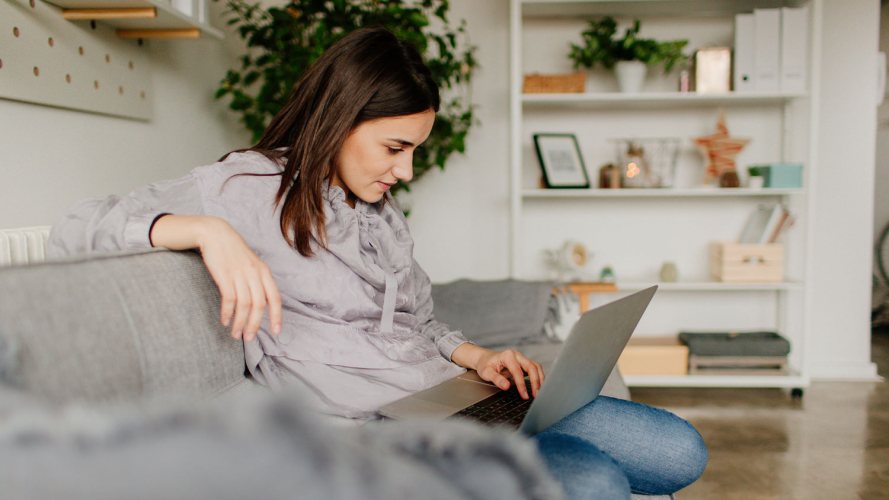 Woman in a sweatshirt, sitting on a grey couch, checking her email on her laptop / consolidate email platforms