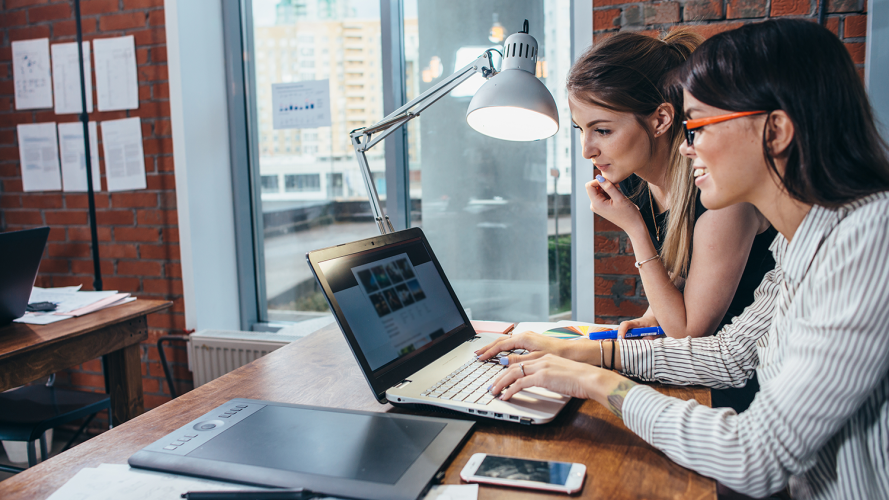 Side profile view of two women collaborating and looking at a laptop screen in an urban office setting.