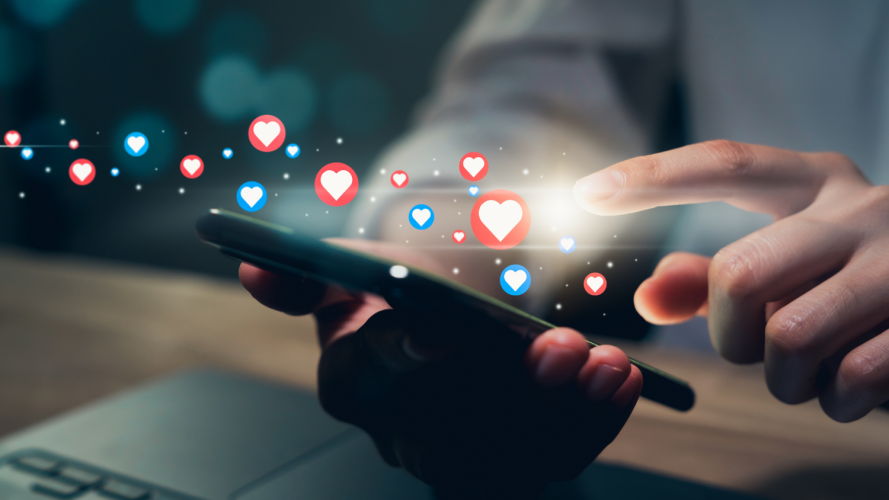 Image shows a pair of hands holding and tapping a mobile phone. There are many heart emoji floating above the screen.