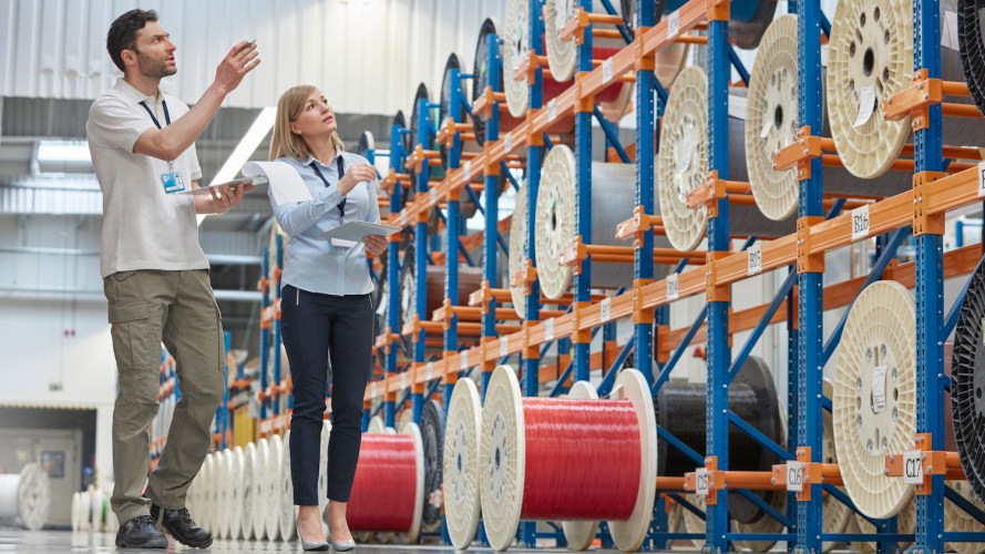 Two warehouse workers, a man and a woman, look over many large spools of fiber. / fiber to the home