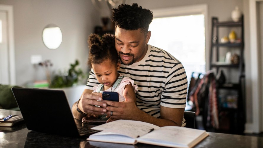 Father sitting at a desk at home, holding his toddler daughter while he checks his phone / customer experience