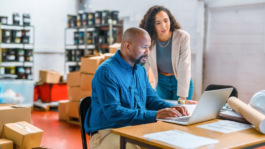 Two coworkers, a man and a woman, sit at a table in a warehouse, looking at a laptop / large language models