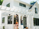 A white house with a pergola and two window installers framing a window.