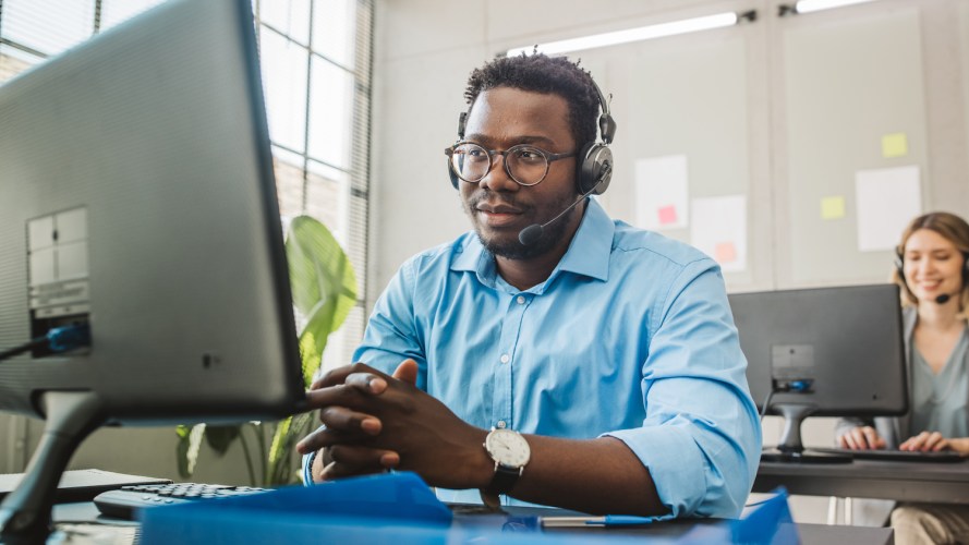 Male office worker, in a blue dress shirt, talking with a customer on headset while looking at his computer in an office setting / contact center automation