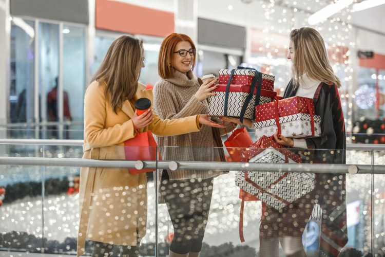 Three women stand in a mall holding wrapped boxes. Holiday shopping predictions say it’s likely they paid for them with credit cards.