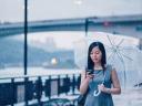 Photograph of a young businesswoman, dressed in a gray dress and holding a clear umbrella, checking her phone on a rainy, overcast day. / drip marketing
