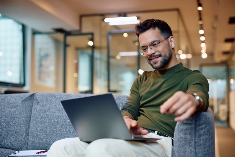 Photo of a young man sitting on a dark blue couch in a modern office setting, checking his laptop / opt-in email marketing