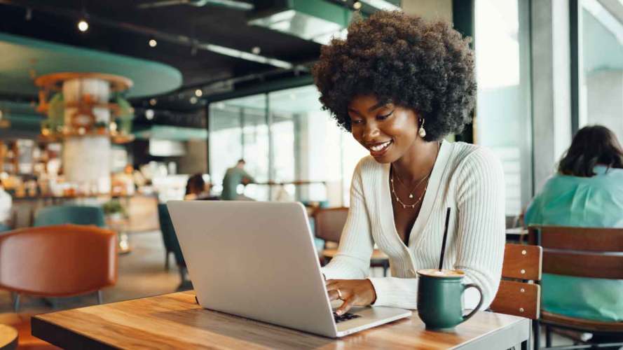 A smiling young Black woman uses her laptop to get self-service customer service in a cafe