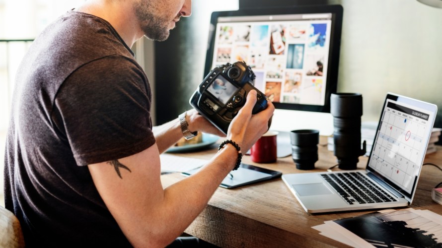 A photographer looks at his camera in front of two computer screens showing video editing software.