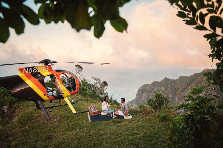 Helicopter landed on a scenic hilltop at sunset, with a couple enjoying a picnic against a mountain background