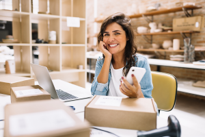 Vendedora sorri enquanto usa um smartphone e um notebook.