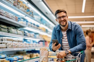 Consumidor compra em supermercado enquanto acessa o smartphone.