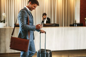 Homem de terno, na recepção do hotel, puxando sua mala e interagindo com o celular.