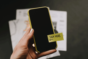 Eine Hand hält ein Smartphone auf dem ein Post-it mit Sign here