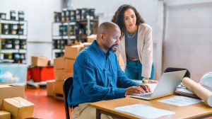 Zwei Mitarbeiter, ein Mann und eine Frau, sitzen an einem Tisch in einer Lagerhalle und schauen auf einen Laptop / große Sprachmodelle