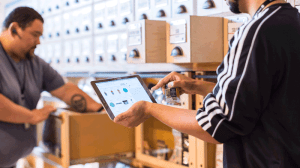 Zwei männliche Personen stehen in einem Lager. Eine Person hält ein Tablet in der Hand.