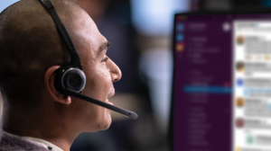 Un hombre con auriculares habla delante de un monitor en el que se ve Slack en el fondo.