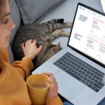 Image of a woman petting a cat and holding a mug while looking at a customer portal on her laptop.