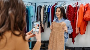 Two women shop for clothing in a store. One woman is taking a photo of her friend, who is holding up a light brown dress. / referral marketing