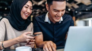 Una pareja joven sonríe mientras trabaja o navega en una computadora portátil. La mujer, que lleva un hijab oscuro y una blusa beige, sostiene un teléfono en una mano y observa la pantalla con una expresión alegre. El hombre, vestido con una camisa azul oscuro, también sonríe mientras escribe en la laptop. Ambos parecen estar disfrutando de la actividad, mostrando una actitud relajada y colaborativa en un ambiente moderno. Esta imagen refleja el trabajo en equipo y la conexión a través de la tecnología.