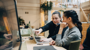 Two young businesswomen sitting at a desk and looking at a computer. The woman on the left is pointing at the computer screen.