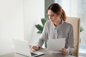 Een gefocuste zakenvrouw typt op een laptop terwijl ze papieren vasthoudt, een rapport voorbereidt en werkresultaten analyseert.